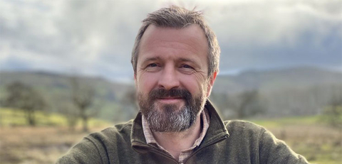 A man with a beard, wearing a zip-up fleece, leans on a gate in front of a field. 