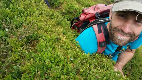 A man in a cap and carrying a rucksack crouches next to a bush, smiling. 