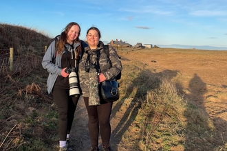 Two young women standing on beach with long-lens cameras and shadows behind  them