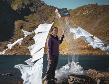 Amy Bray standing next to her Another Way sculpture in the mountains, with a lake behind her.