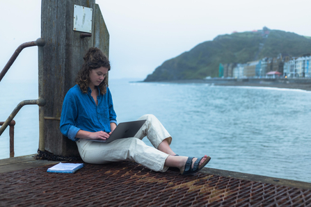 a woman wearing blue top and white trousers, sat by the sea using a laptop